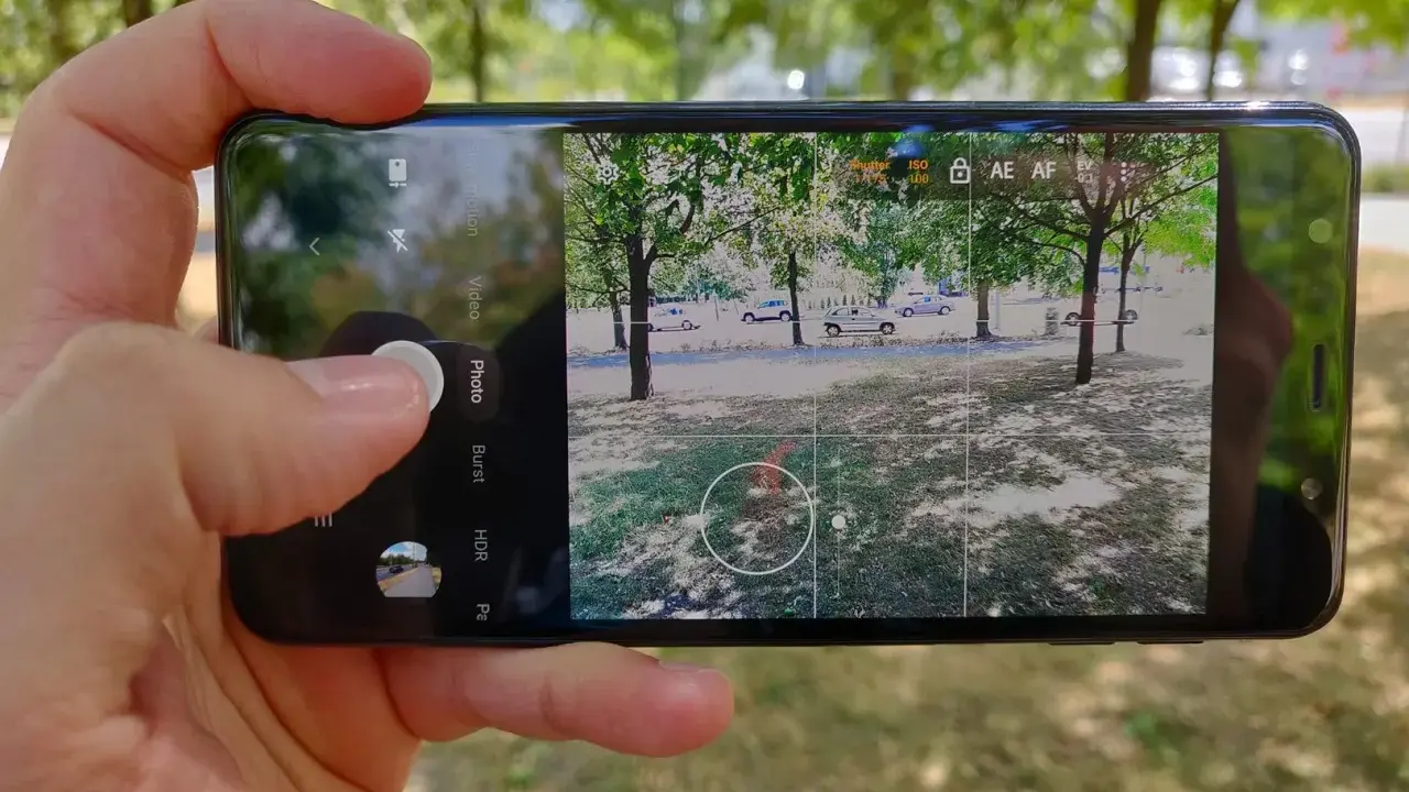 Dłoń trzyma smartfon z otwartą aplikacją do robienia zdjęć. Widok na park z drzewami i samochodami.