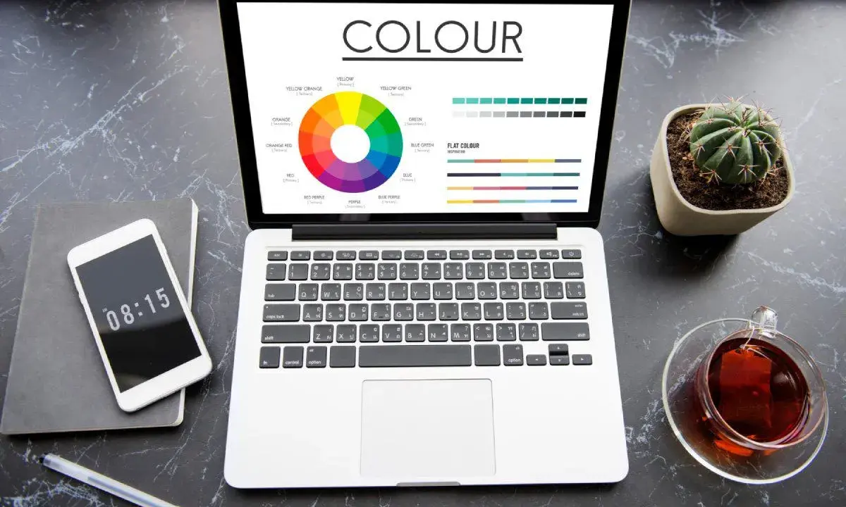 Na ekranie laptopa widać koło barw i wykresy, porównujące modele kolorów rgb vs cmyk. Obok telefon, notes i filiżanka herbaty.