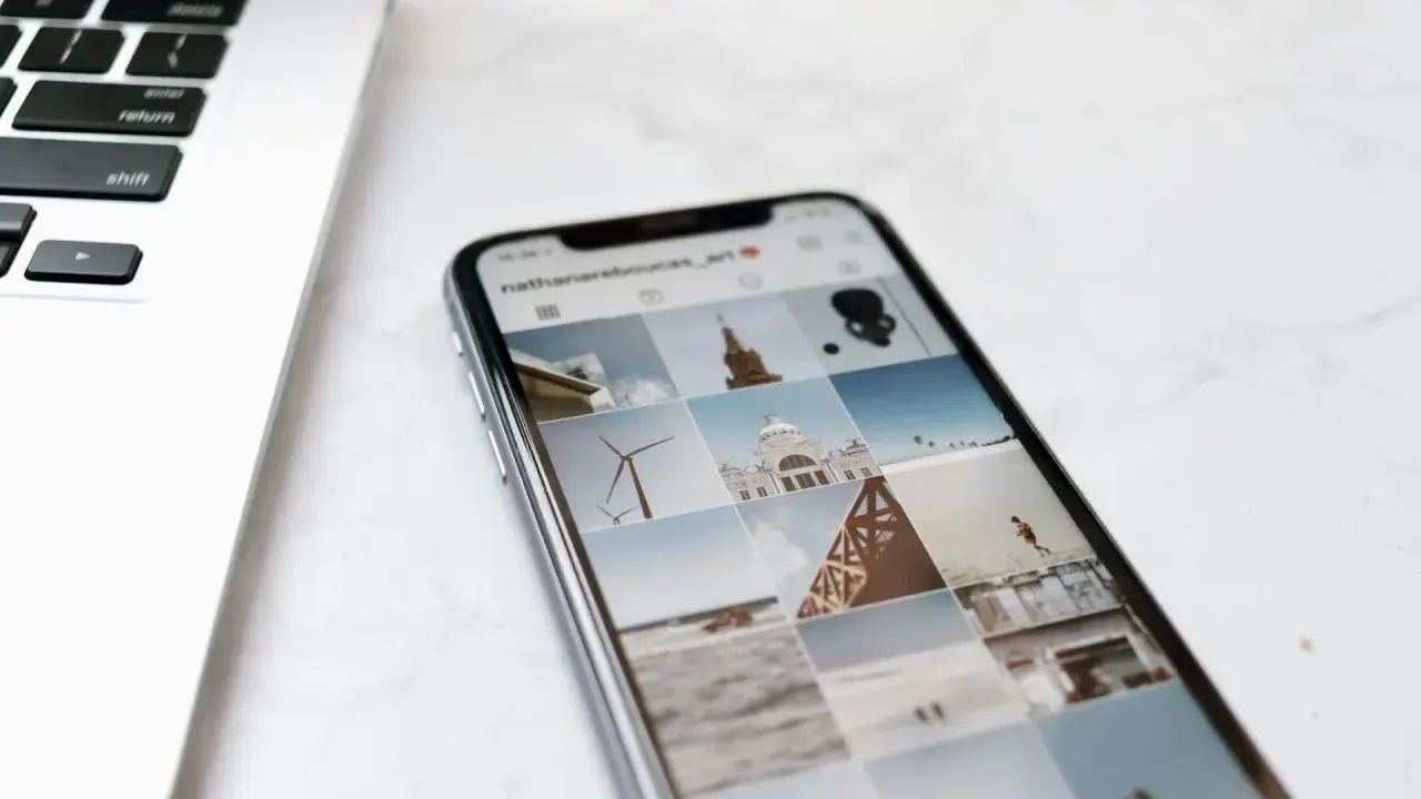 Telefon z Instagramem obok laptopa. Na ekranie telefonu widać siatkę zdjęć, gdzie są aktualności na Instagramie, m.in. wiatrak i budynek.