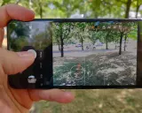 Dłoń trzyma smartfon z otwartą aplikacją do robienia zdjęć. Widok na park z drzewami i samochodami.