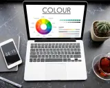 Na ekranie laptopa widać koło barw i wykresy, porównujące modele kolorów rgb vs cmyk. Obok telefon, notes i filiżanka herbaty.