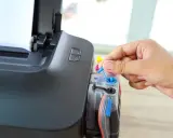 Dłoń zamyka niebieską zatyczkę w drukarce Epson, przygotowując ją do połączenia z laptopem przez Wi-Fi.