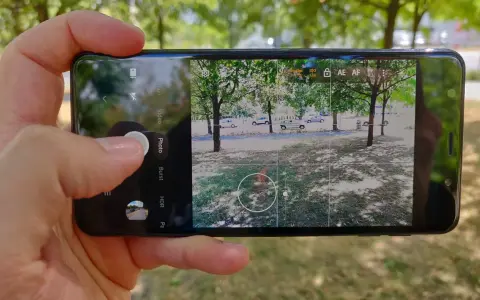 Dłoń trzyma smartfon z otwartą aplikacją do robienia zdjęć. Widok na park z drzewami i samochodami.