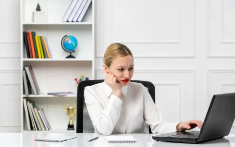 Zamyślona kobieta przy laptopie, być może przegląda komunikatory firmowe. Na półce za nią stoją książki i globus.