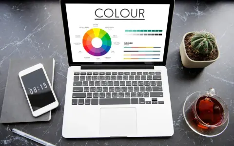 Na ekranie laptopa widać koło barw i wykresy, porównujące modele kolorów rgb vs cmyk. Obok telefon, notes i filiżanka herbaty.