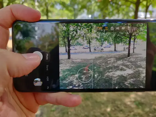 Dłoń trzyma smartfon z otwartą aplikacją do robienia zdjęć. Widok na park z drzewami i samochodami.