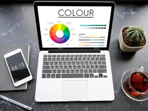 Na ekranie laptopa widać koło barw i wykresy, porównujące modele kolorów rgb vs cmyk. Obok telefon, notes i filiżanka herbaty.