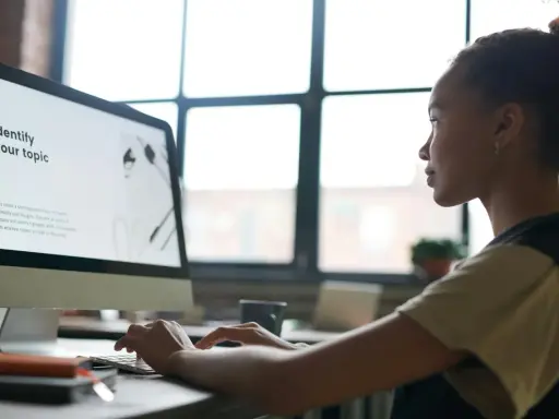 Kobieta pracuje przy komputerze, na ekranie widoczny jest tekst "Identify your topic". Może szukać informacji, jak wstawić tło w Wordzie.