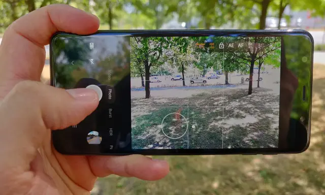 Dłoń trzyma smartfon z otwartą aplikacją do robienia zdjęć. Widok na park z drzewami i samochodami.