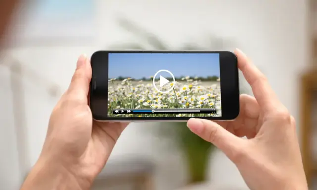 Dłonie trzymają smartfon z widokiem na pole stokrotek. Na ekranie odtwarzacz wideo, idealny do edycji i przycinania filmów.