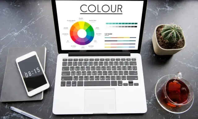 Na ekranie laptopa widać koło barw i wykresy, porównujące modele kolorów rgb vs cmyk. Obok telefon, notes i filiżanka herbaty.