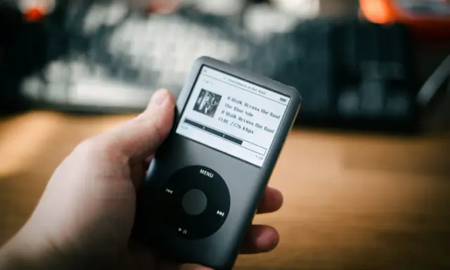 Dłoń trzyma iPoda z listą utworów, w tym "A Walk Across the Roof". Idealny do słuchania muzyki, jakbyś używał audiograbber po polsku.