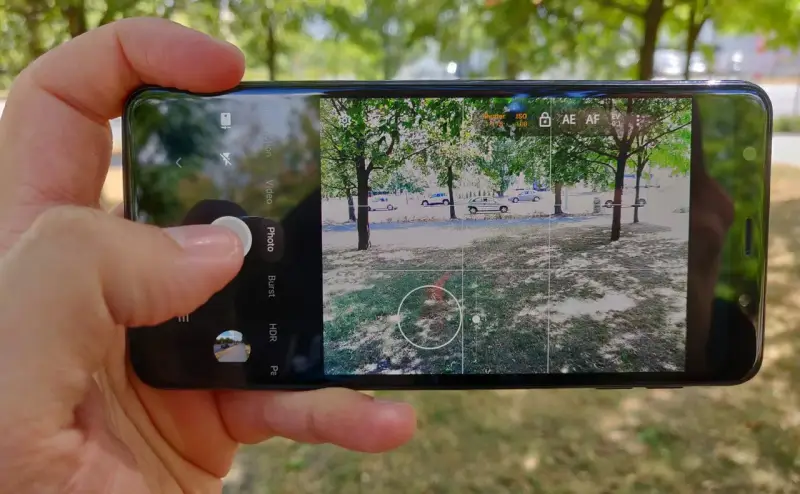 Dłoń trzyma smartfon z otwartą aplikacją do robienia zdjęć. Widok na park z drzewami i samochodami.